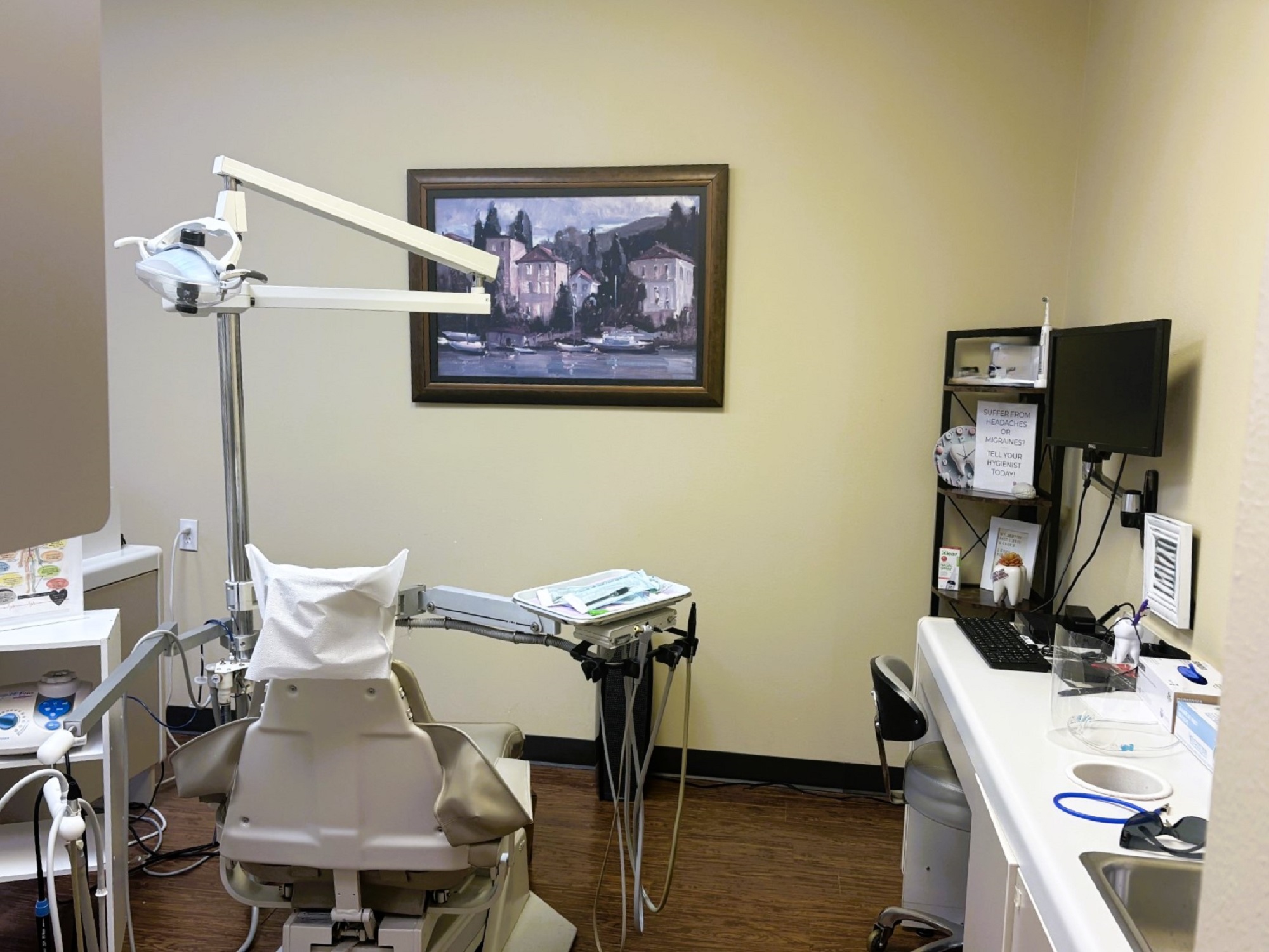 Light tan walls with dark baseboards and dark hardwood flooring. A large framed piece of art hangs on the wall. A tan dental hygiene chair anchors the room with overhead dental lighting. Rolling trays and small cabinets surround the chair with lots of tools, supplies, and equipment. There's a white cabinet on one wall with a small sink and lots of drawers and space for supplies and tools.