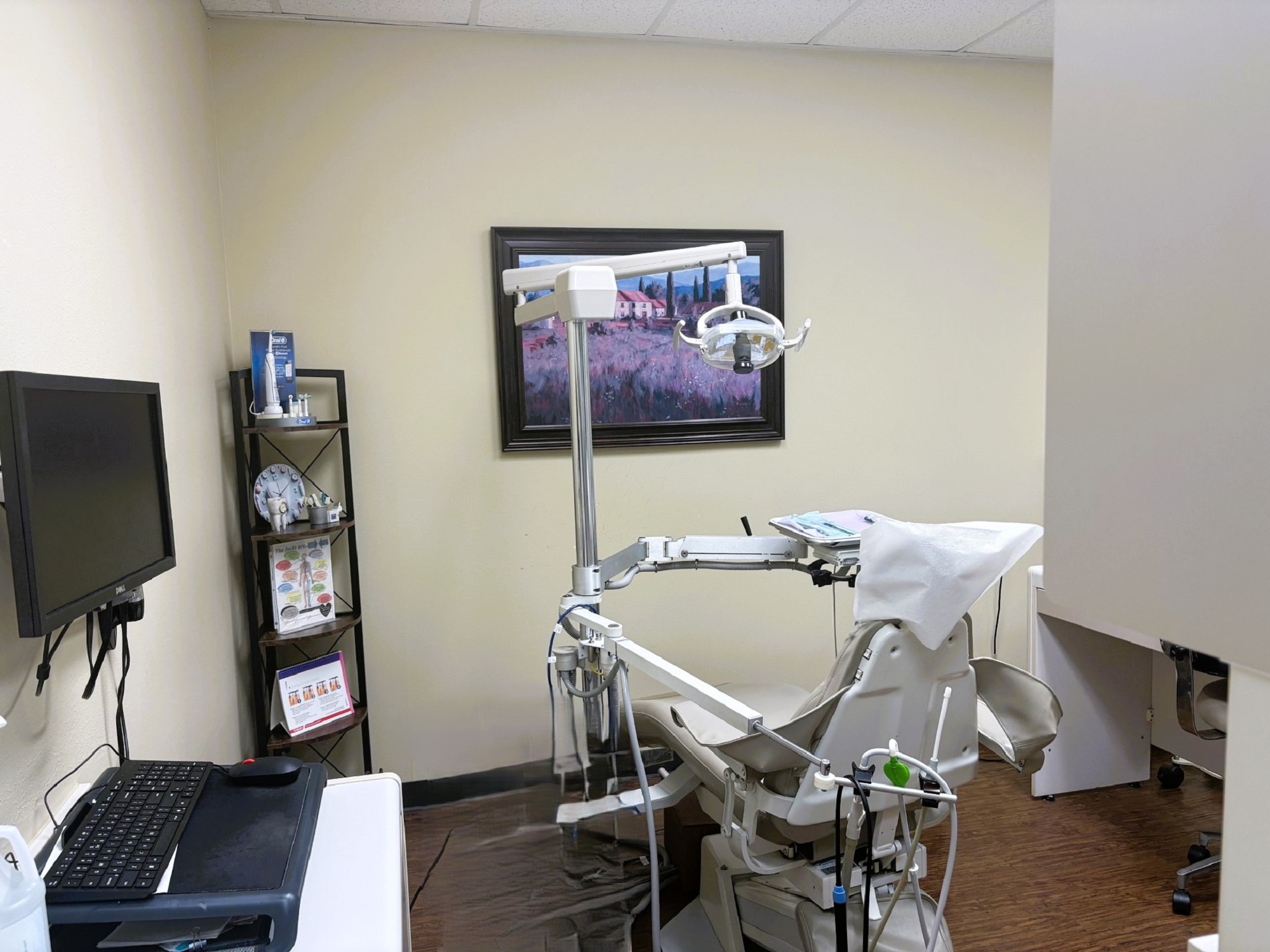 Light tan walls with dark baseboards and dark hardwood flooring. A large framed piece of art hangs on the wall. A tan dental hygiene chair anchors the room with overhead dental lighting. Rolling trays and small cabinets surround the chair with lots of tools, supplies, and equipment. There's a white cabinet on one wall with a small sink and lots of drawers and space for supplies and tools.