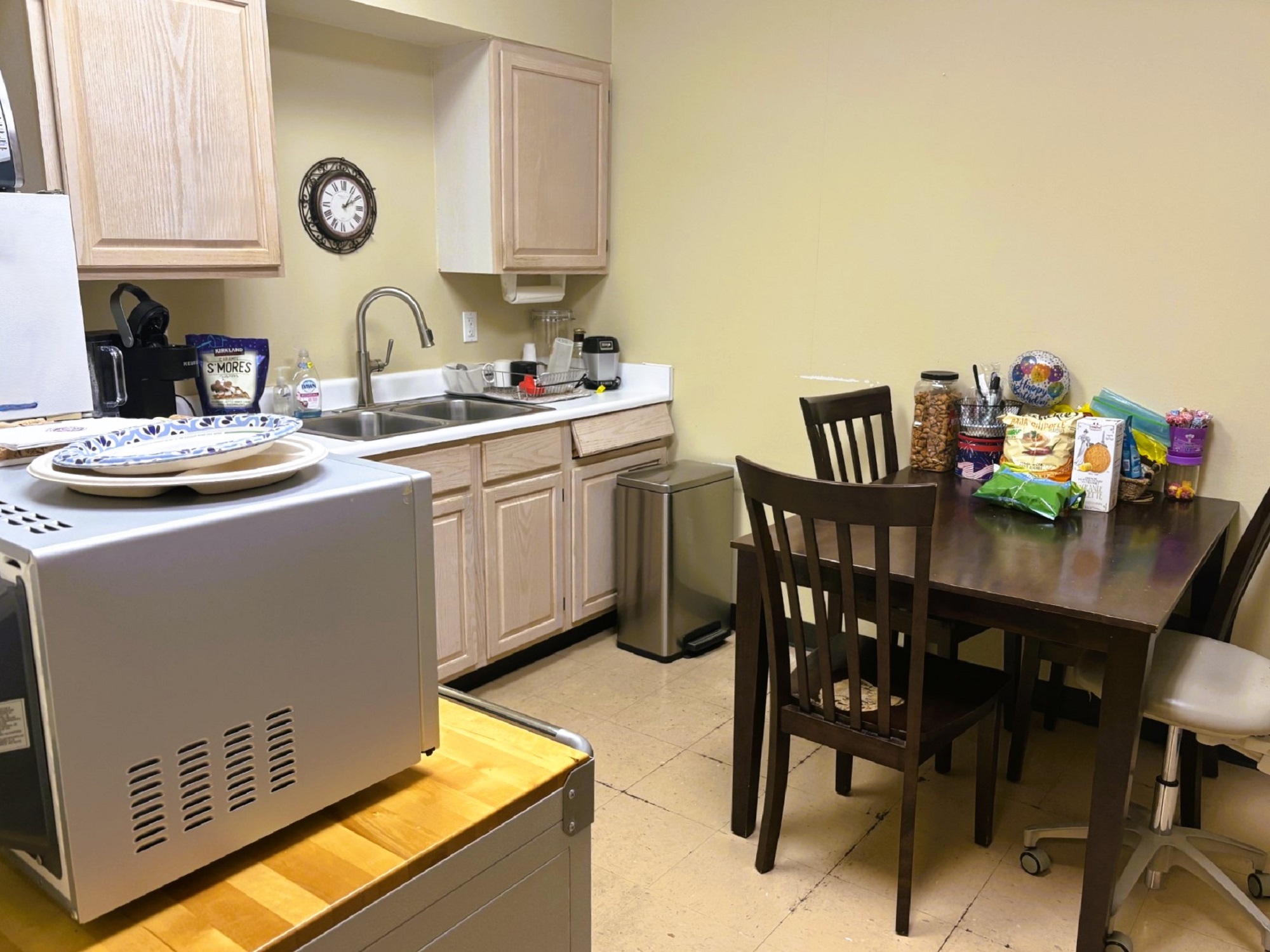 Tan walls with dark baseboards and brown flooring. A small square table sits against one wall with four chairs around it. Bleached wood cabinetry hangs on the wall with white countertops and a stainless steel sink. A microwave sits in one corner next to a refrigerator.