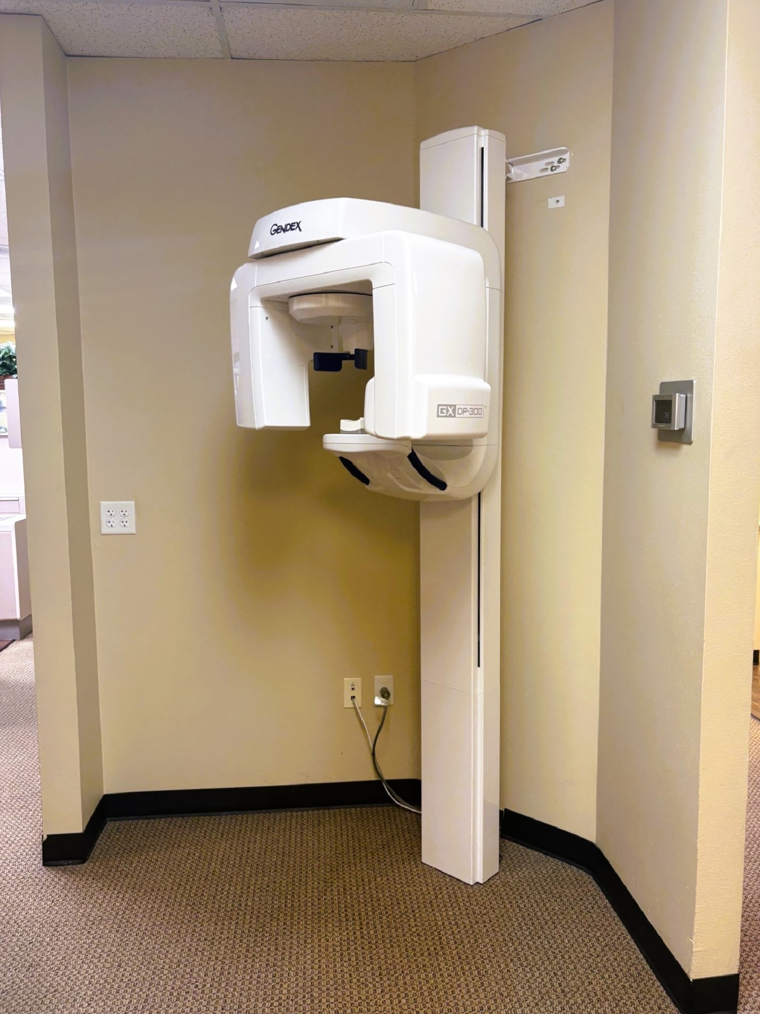 Tan walls with dark baseboards and slightly lighter brown commercial carpet flooring. A large panorex x-ray machine stands in the corner.