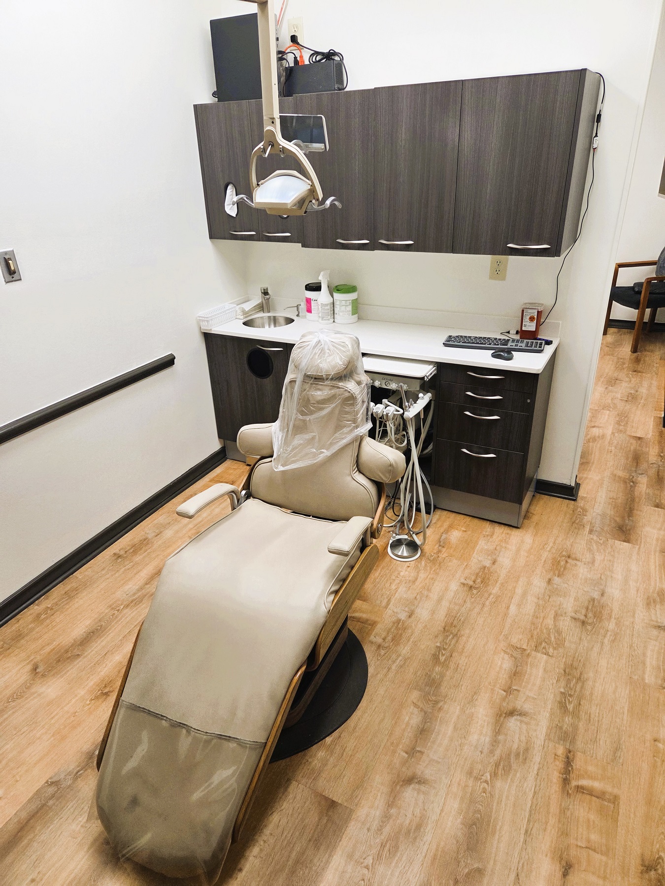 Light walls with dark chair rail and base boards. Light wood look flooring. Light cabinetry behind a light dental procedural chair that anchors the room. There's an overhead dental procedural light. There's both an upper and lower cabinet to hold lots of dental tools, supplies, and equipment.