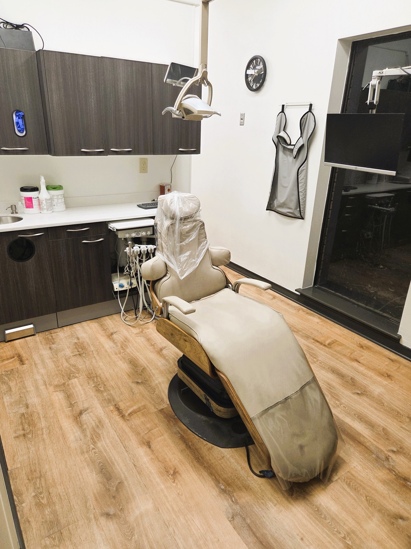 Light walls with dark chair rail and base boards. Light wood look flooring. Light cabinetry behind a light dental procedural chair that anchors the room. There's an overhead dental procedural light. There's both an upper and lower cabinet to hold lots of dental tools, supplies, and equipment.