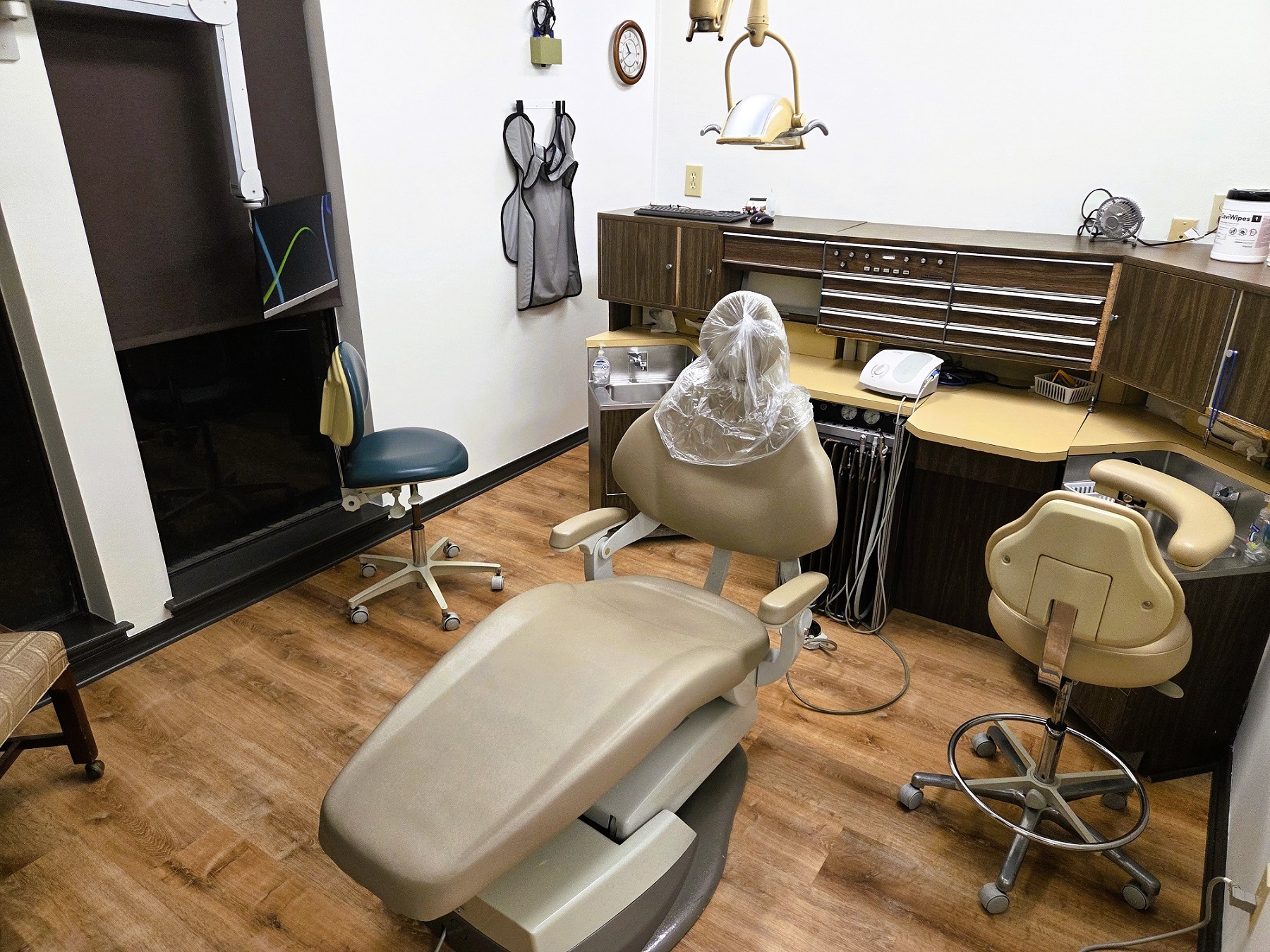 Light walls with dark chair rail and base boards. Light wood look flooring. Light cabinetry behind a light dental procedural chair that anchors the room. There's an overhead dental procedural light. There's both an upper and lower cabinet to hold lots of dental tools, supplies, and equipment.