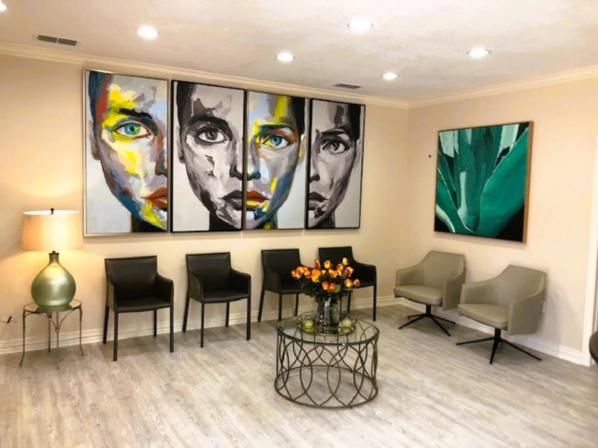 Beige walls with cream crown molding and tall base boards. Grey stained wood look flooring. Multiple can lights light up the over sized artwork on the walls. Several chairs sit along the wall under the art with an end table holding a large lamp. There's a round coffee table sitting in the middle of the room.