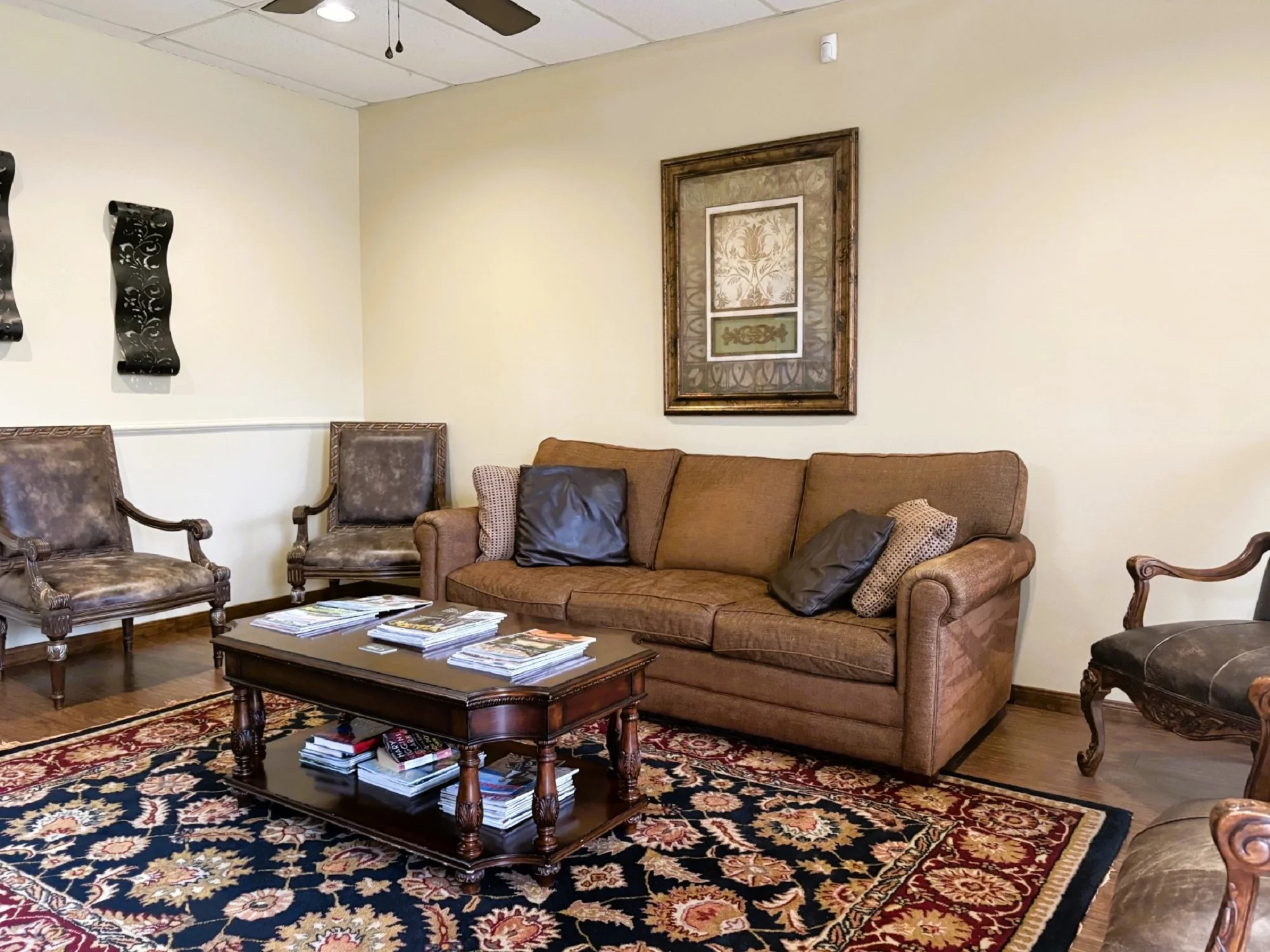 Light walls with matching chair rail and dark baseboards and matching dark hardwood flooring. A huge area rug covers most of the hardwood flooring with a brown overstuffed couch on one wall sitting under a large framed piece of art. Brown leather arm chairs surround the room sitting on the wall with other artwork above. A dark coffee table anchors the room with lots of magazines.