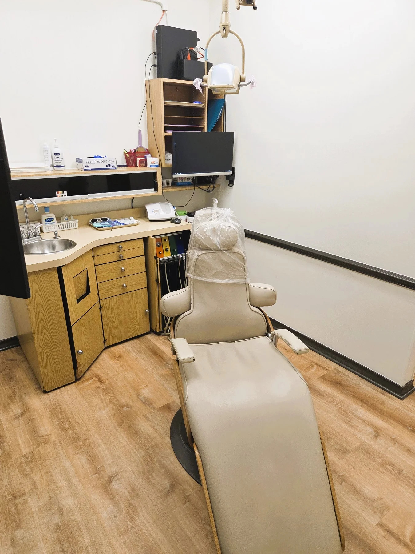 Light walls with dark chair rail and base boards. Light wood look flooring. Light cabinetry behind a light dental procedural chair that anchors the room. There's an overhead dental procedural light. There's both an upper and lower cabinet to hold lots of dental tools, supplies, and equipment.